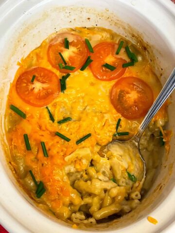 A slow cooker pot with cooked macaroni cheese with sliced tomatoes and chopped chives with serving spoon in the dish.