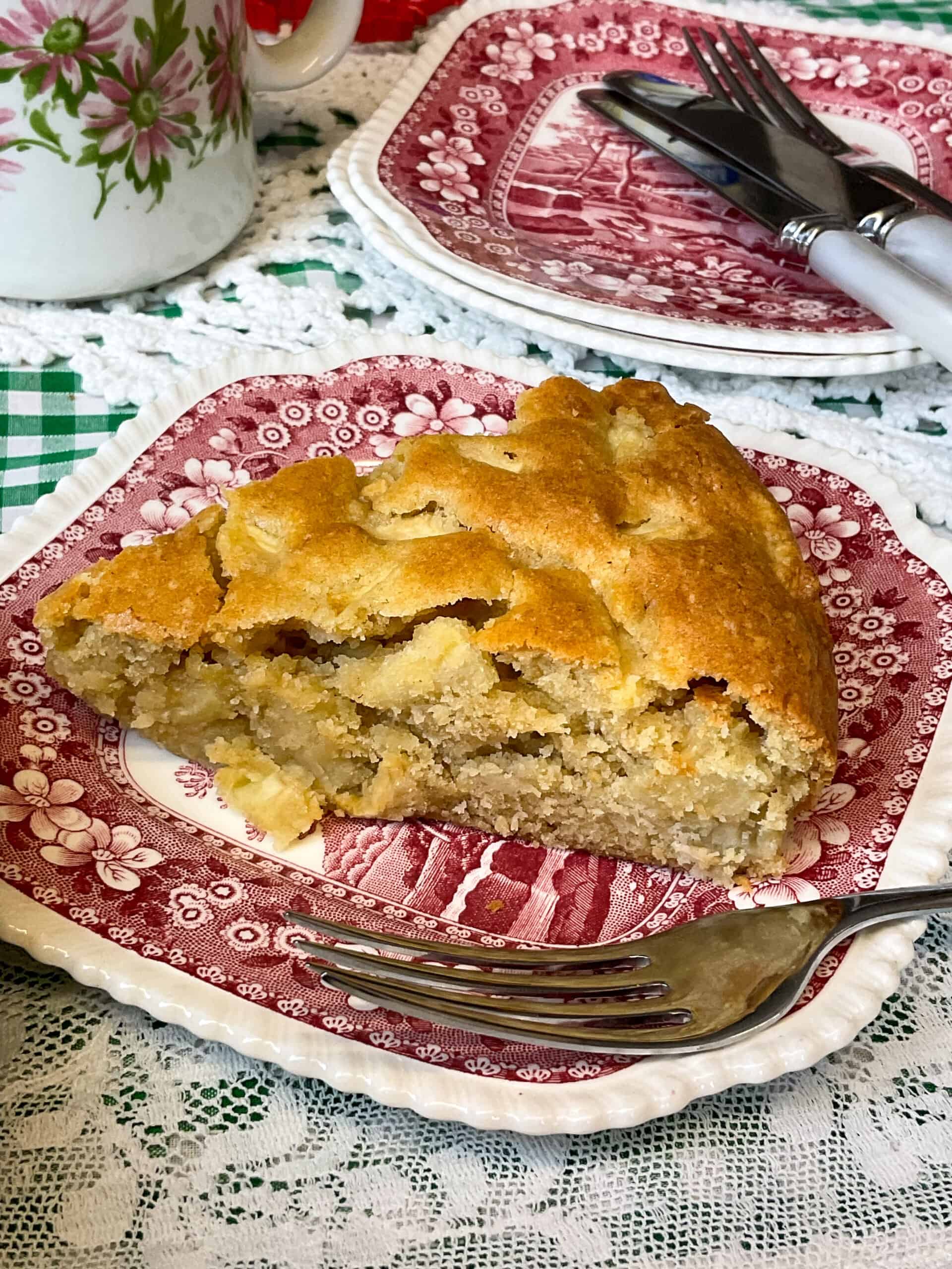 Two small vintage red flower patterned cake plates with one plate having a slice of apple cake and a cake fork, flower patterned cream jug to side, and a white doily tablecloth.