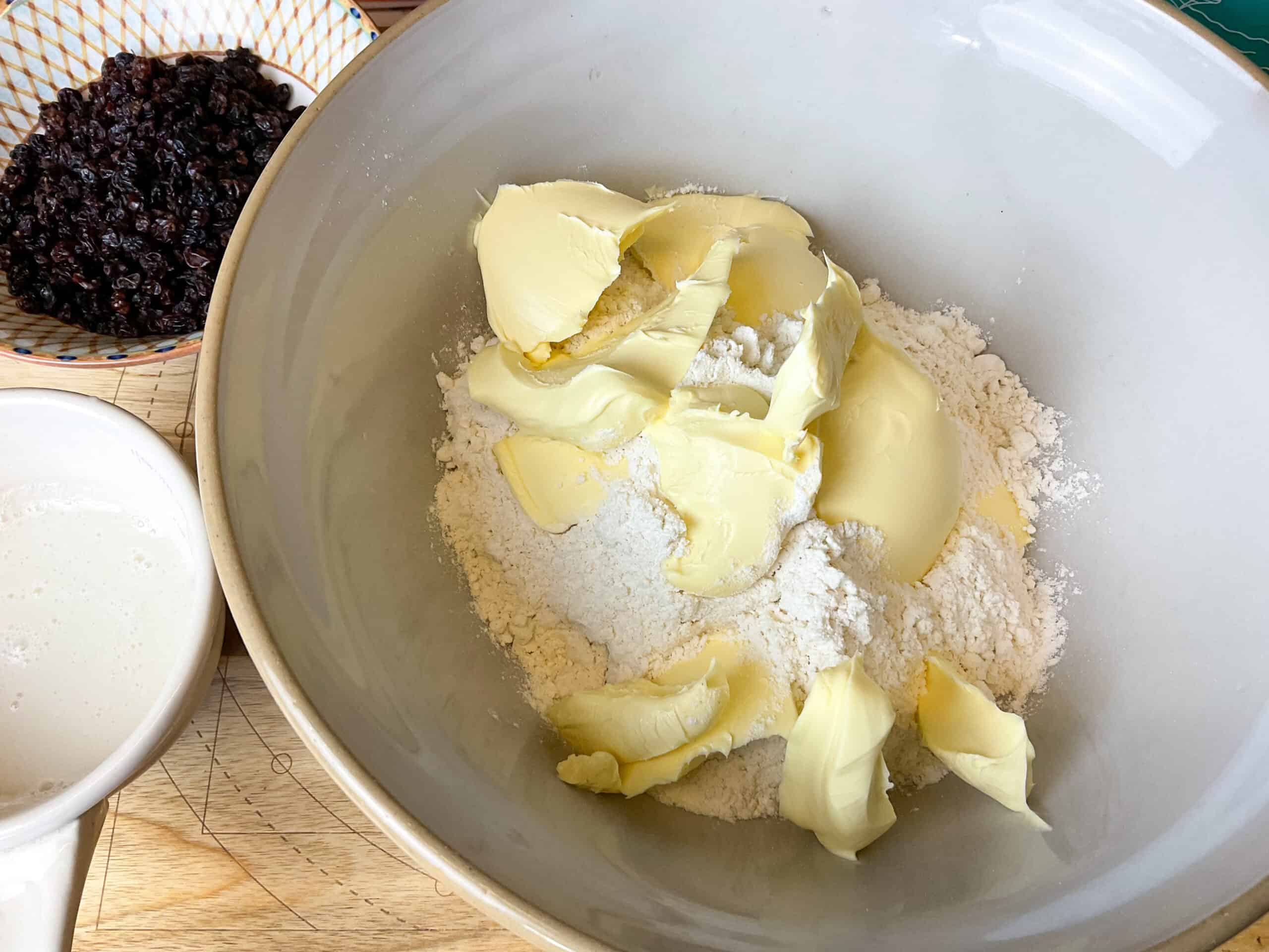 Flour and margarine in mixing bowl with small bowl of currants to side and small dish of milk.