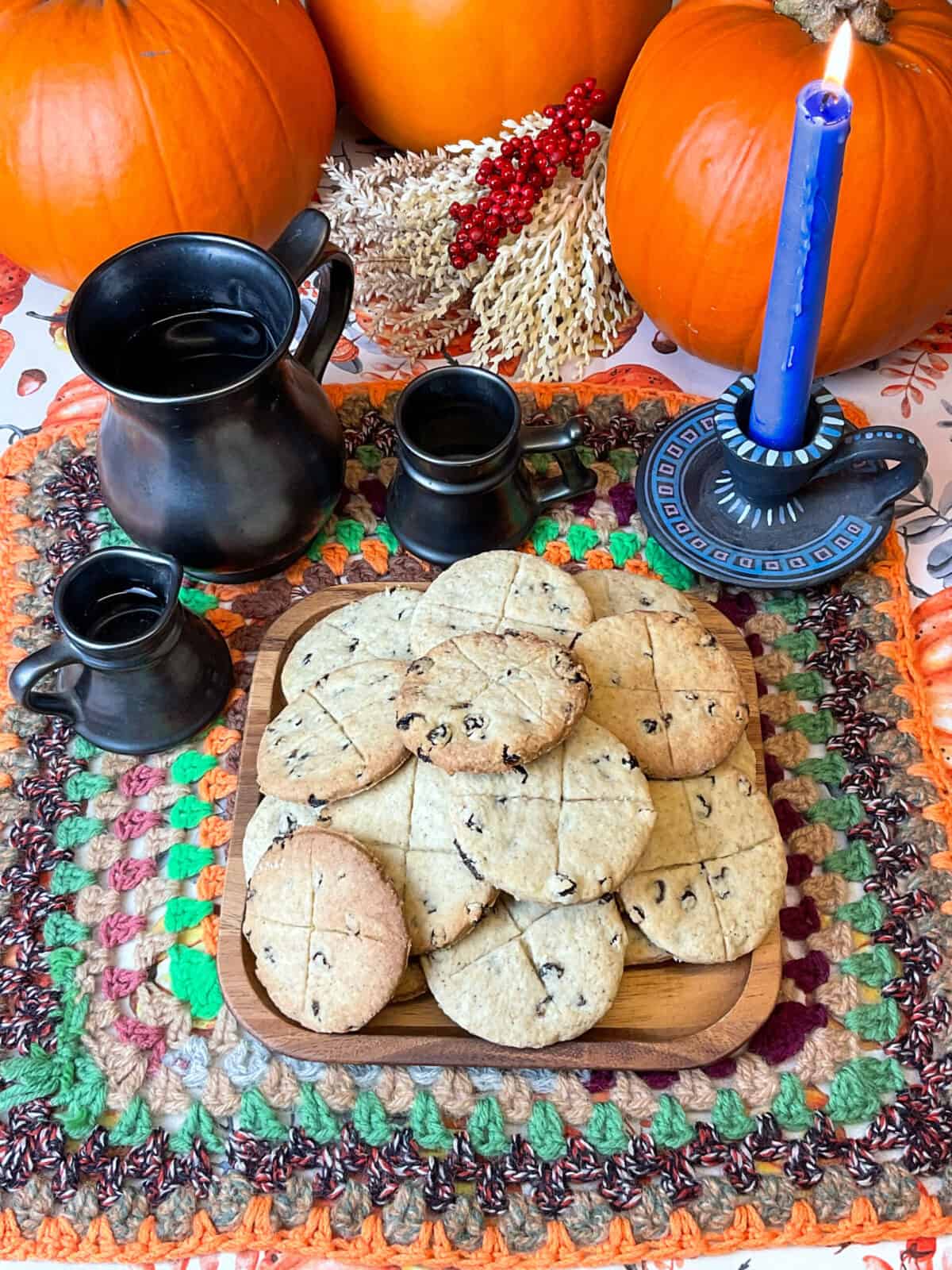 A plate of soul cakes with a blue candle lit and pewter cups, jugs, in background, and a crochet multi-coloured table mat and pumpkin background.