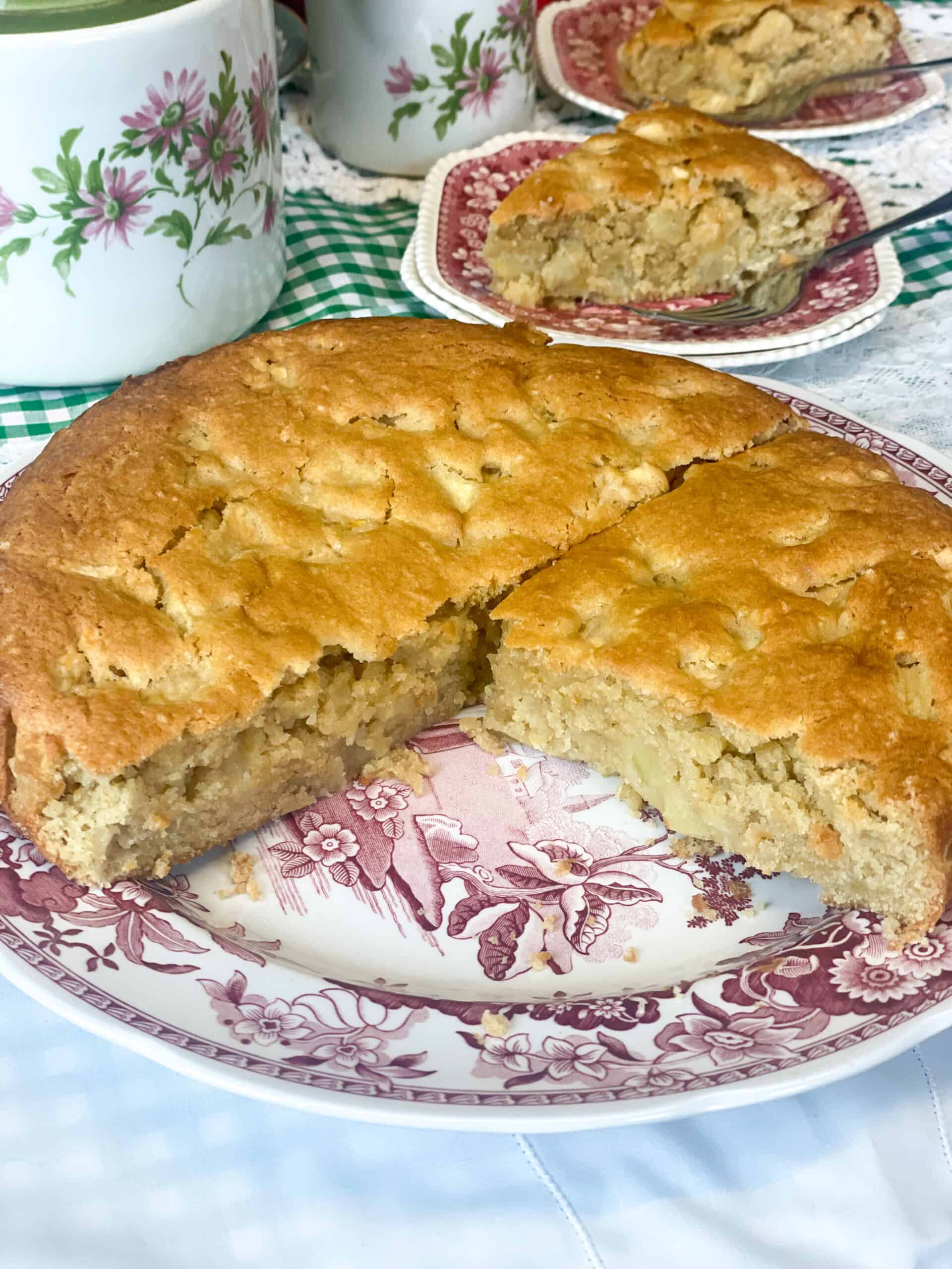 Apple cake on cake plate and sliced and pieces of cake on small dessert plates with flower patterned teapot in background.