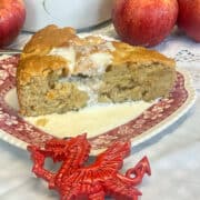 Slice of Welsh apple cake with cream poured over, small Welsh dragon ornament in front, apples in background with a teapot, featured image.