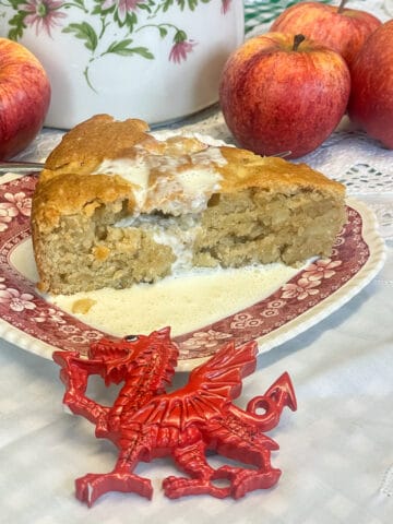 Slice of Welsh apple cake with cream poured over, small Welsh dragon ornament in front, apples in background with a teapot, featured image.