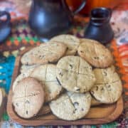 A wooden square plate with pile of soul cakes sitting on a crochet table mat with pewter pitcher and mug in background along with pumpkins.