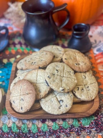 A wooden square plate with pile of soul cakes sitting on a crochet table mat with pewter pitcher and mug in background along with pumpkins.