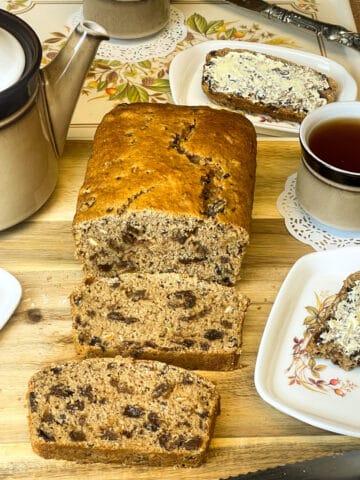 Bara Birth on cutting board with three slices cut, brown teapot to side and matching cup to side, buttered slice of tea bread in background and a small flower imaged tea tray to side with slice of fruit bread.