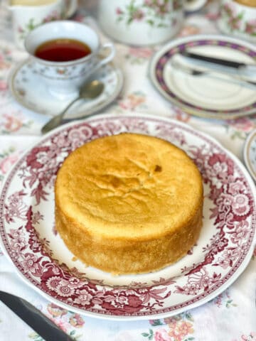 A ground rice cake on a red patterned vintage cake plate, with vintage tea cup and saucer, and serving plate in background, pink and white flowered table cloth.
