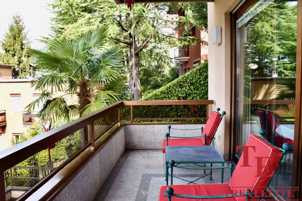 Luxurious Italian balcony with elegant red and teal outdoor furniture overlooking lush greenery and modern architecture. Perfect for relaxing in style amidst nature.