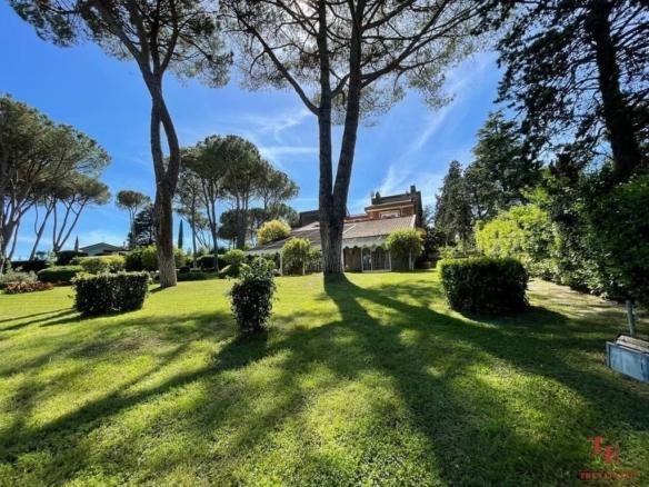 Lush green garden with tall trees and a luxurious villa in the background under a bright blue sky, showcasing premier Italian real estate elegance.