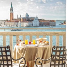View of a Venetian cityscape through an open balcony door with a breakfast table set for two with orange juice and pastries, overlooking a canal and historic buildings in Italy.