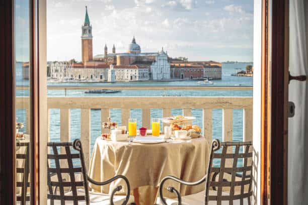 View of a Venetian cityscape through an open balcony door with a breakfast table set for two with orange juice and pastries, overlooking a canal and historic buildings in Italy.