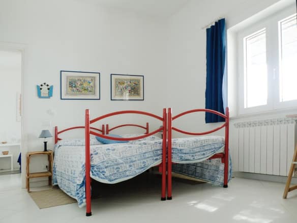 Bright bedroom with red metal bed frame, blue curtains, colourful wall art, and white walls, showcasing a charming Italian property by Trevi Elite.