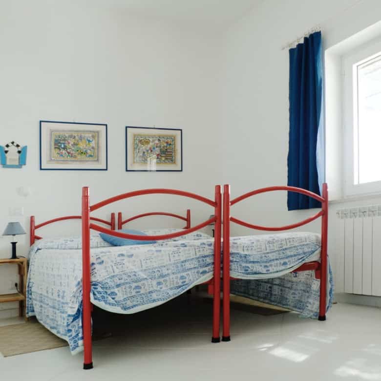 Bright bedroom with red metal bed frame, blue curtains, colourful wall art, and white walls, showcasing a charming Italian property by Trevi Elite.