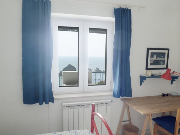 Bright room with a view of the sea and rocky coast, featuring a large window with blue curtains, a wooden desk, colourful decor, and framed photos, showcasing a cosy Italian holiday apartment.