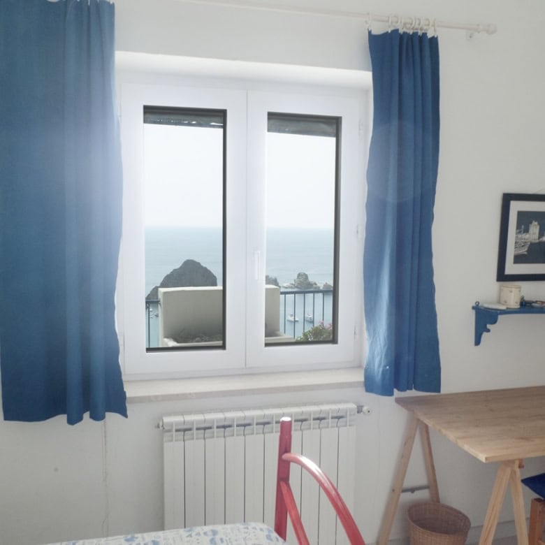 Bright room with a view of the sea and rocky coast, featuring a large window with blue curtains, a wooden desk, colourful decor, and framed photos, showcasing a cosy Italian holiday apartment.