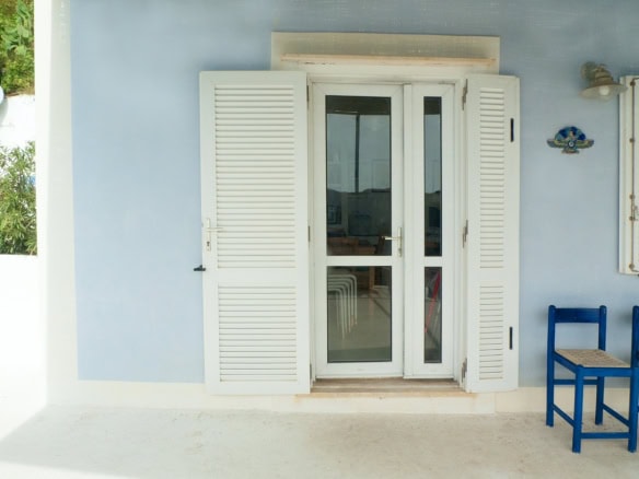 Bright blue window shutters and a glass door on a pastel blue Mediterranean-style building, with a small outdoor seating area featuring blue chairs. Perfect for a seaside villa or coastal holiday home in Italy.