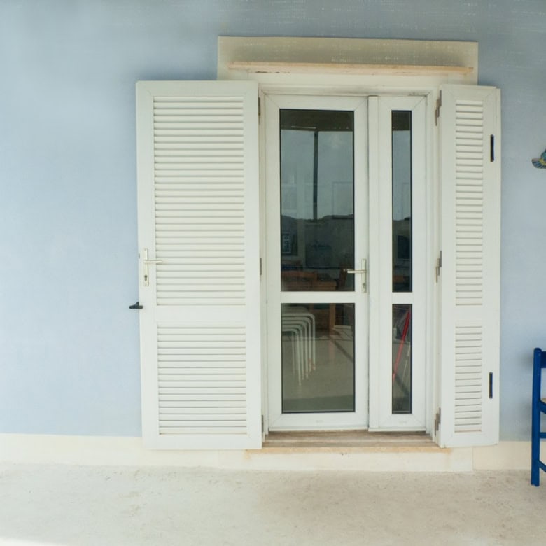 Bright blue window shutters and a glass door on a pastel blue Mediterranean-style building, with a small outdoor seating area featuring blue chairs. Perfect for a seaside villa or coastal holiday home in Italy.