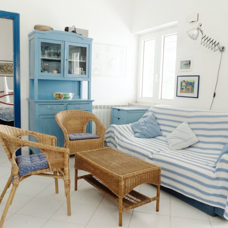 Bright living room featuring a blue and white striped sofa, wicker chairs, and a vintage blue cabinet, offering a coastal-inspired Italian real estate aesthetic.