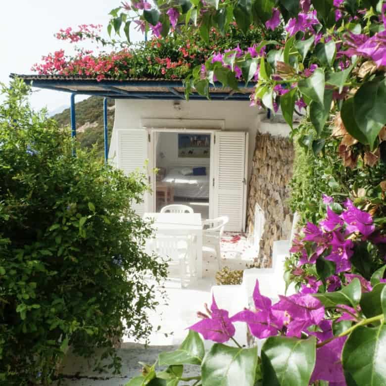 Rose-covered terrace leading to a charming Italian villa with white shutters and lush greenery in a picturesque countryside setting.