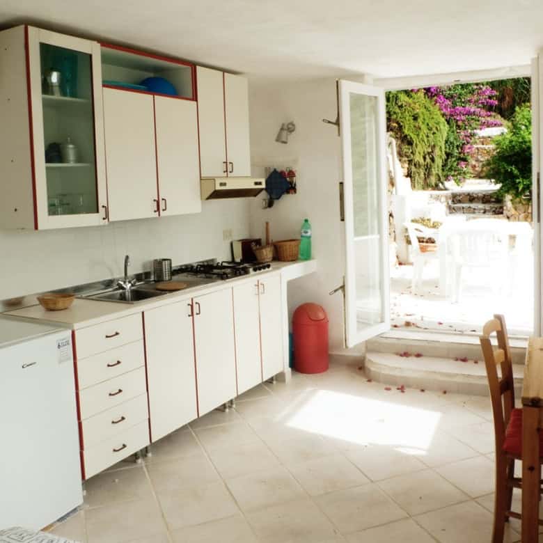Bright and airy Italian kitchen with outdoor access, featuring white cabinets with red trim, a small refrigerator, wooden dining table, and views of colourful Mediterranean garden and stone steps.