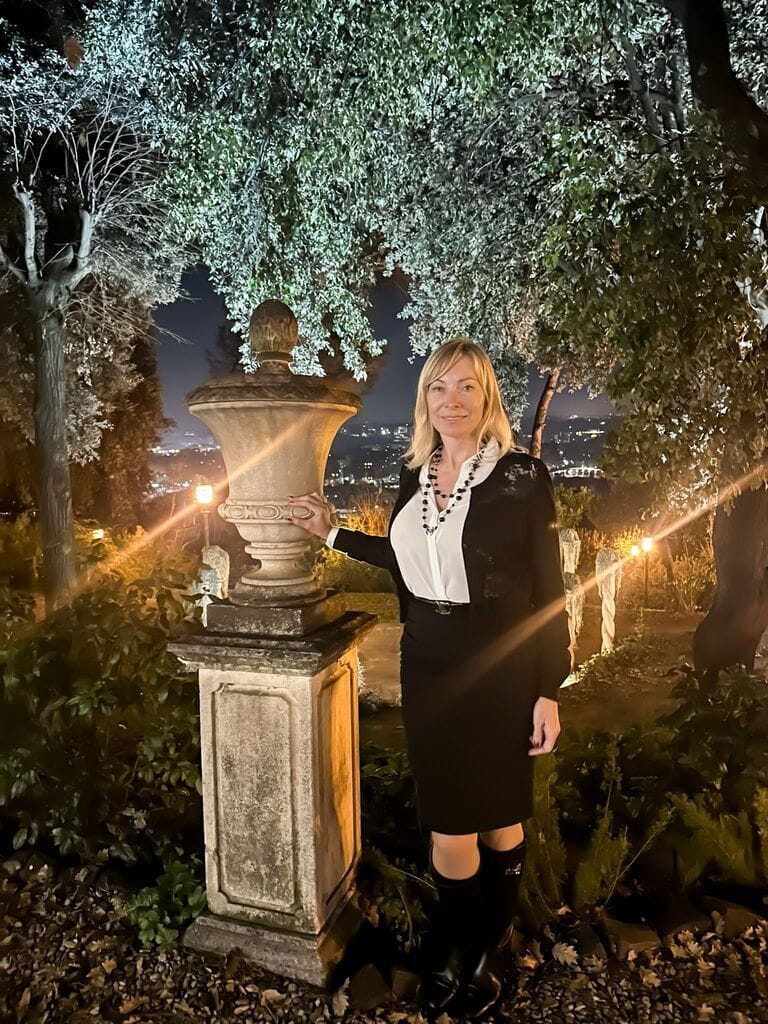 Elegant woman in black and white outfit standing beside a classical stone urn in a beautifully lit Italian garden at night, with city lights in the background.