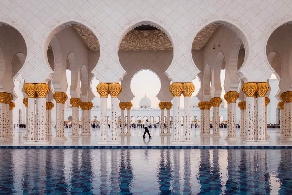 Luxurious mosque architecture with ornate white columns and gold accents, reflecting in a serene pool, showcasing Islamic art and design elements in a grand setting.