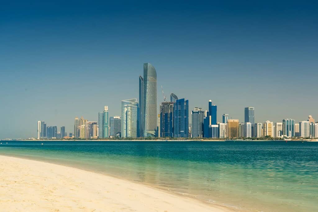 Stunning Dubai city skyline with modern skyscrapers along the waterfront, viewed from a sandy beach beneath a clear blue sky. An ideal setting for luxury real estate investments.