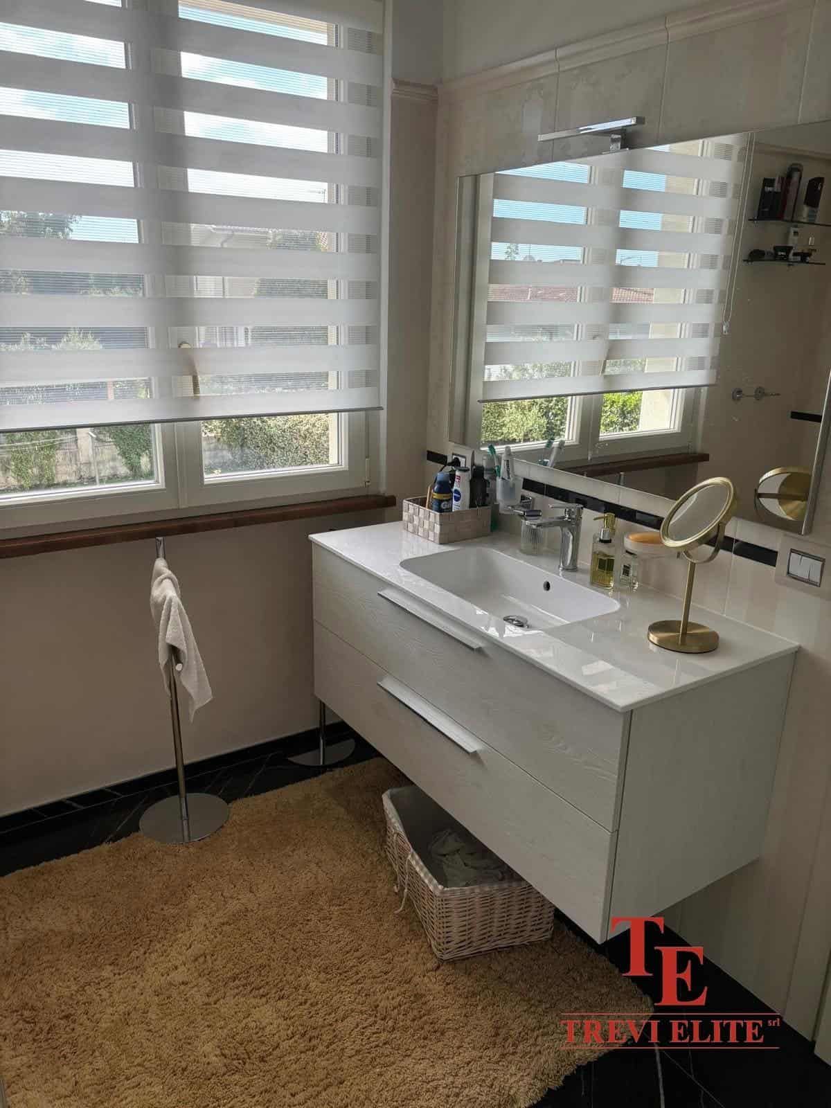 Bright modern bathroom with white vanity unit, large mirror, and double windows with striped blinds, natural light, and minimal decor. Perfect for a stylish Italian property.