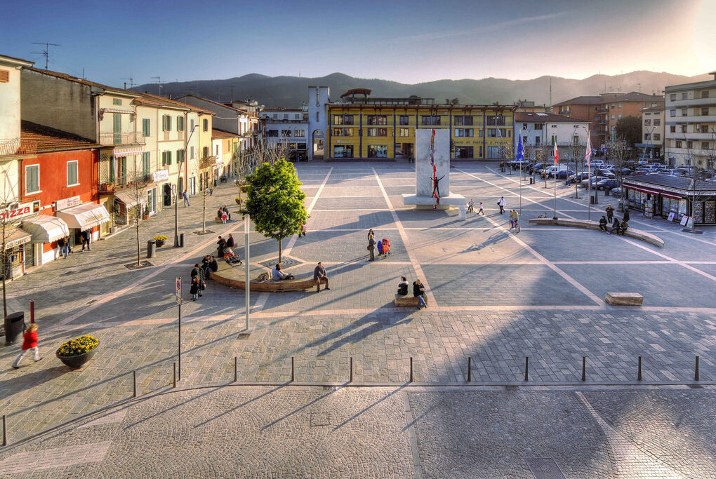 Ancient Italian town square with colourful historic buildings, modern benches, and a prominent monument, set against a backdrop of mountains at sunset for luxury connoisseurs - Trevi Elite.