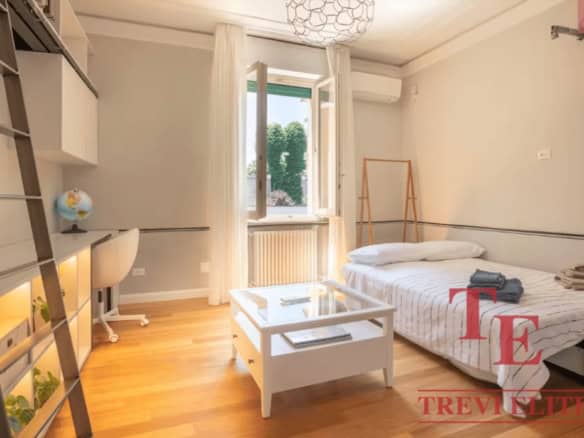 Bright bedroom with natural light, white decor, wooden flooring, large window with curtains, study desk, and a cozy bed, showcasing stylish Italian real estate options by Trevi Elite.