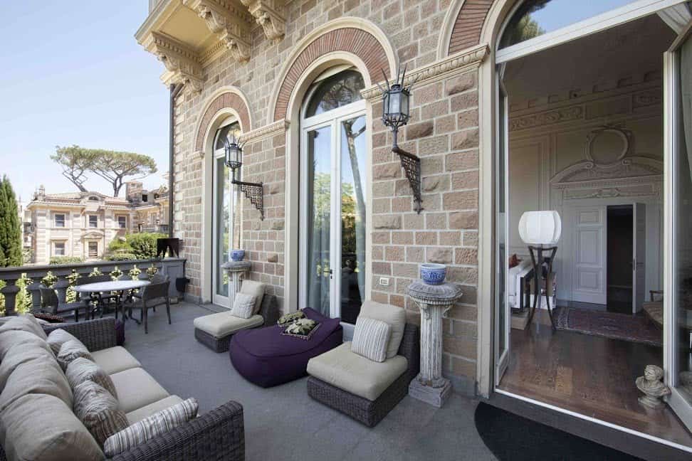 Luxurious Italian balcony featuring classic architecture, elegant wicker furniture, ornate wall sconces, and scenic views of historic buildings and lush greenery in Italy.
