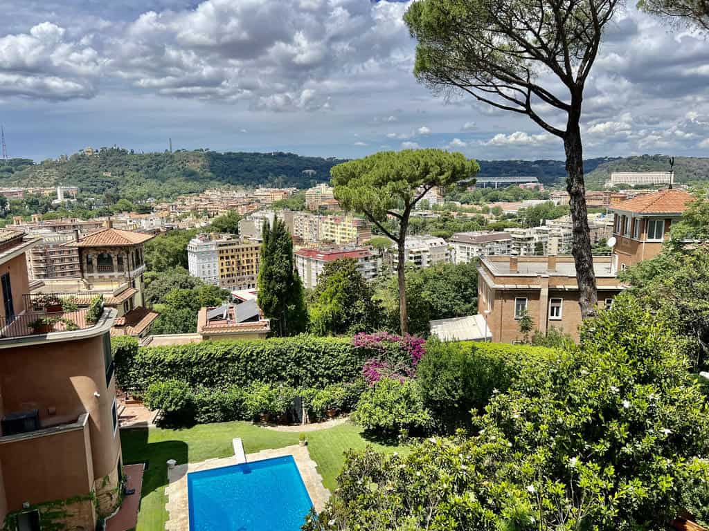 Stunning view of a lush Italian hillside neighbourhood with a private pool, vibrant greenery, and picturesque cityscape in the background under a partly cloudy sky.