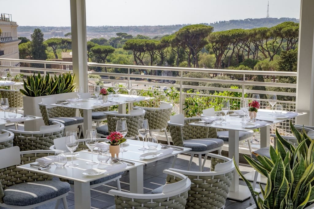 Elegant outdoor restaurant terrace with stylish white tables, comfortable woven chairs, and vibrant floral centrepieces, offering panoramic views of Rome’s lush greenery and iconic skyline.