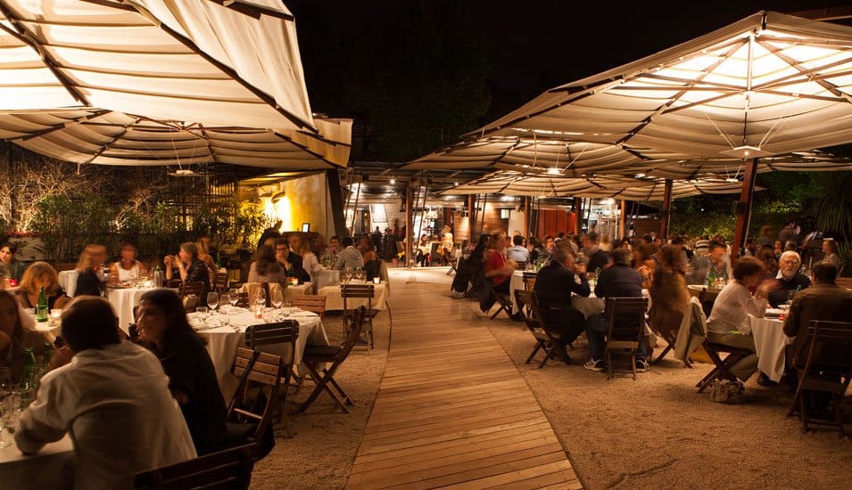 Luxurious outdoor Italian restaurant at night with elegantly dressed diners, ambient lighting, under large canopy umbrellas, offering an exclusive dining experience in Italy.