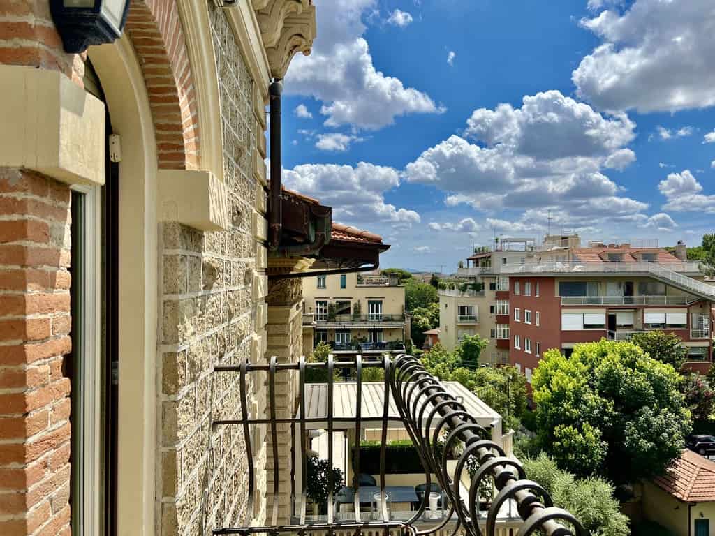 Charming balcony view overlooking lush greenery and modern apartment buildings under a bright blue sky with fluffy clouds in Italy. Perfect for luxury property listings.