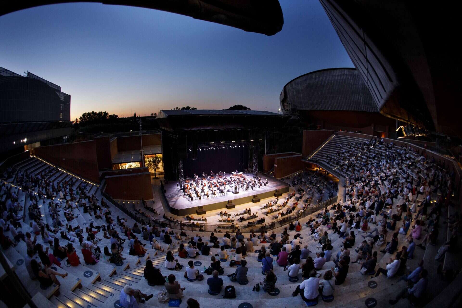 Vibrant outdoor concert at dusk in a modern Italian theatre with an audience enjoying live music and stage performances amid stylish architecture.