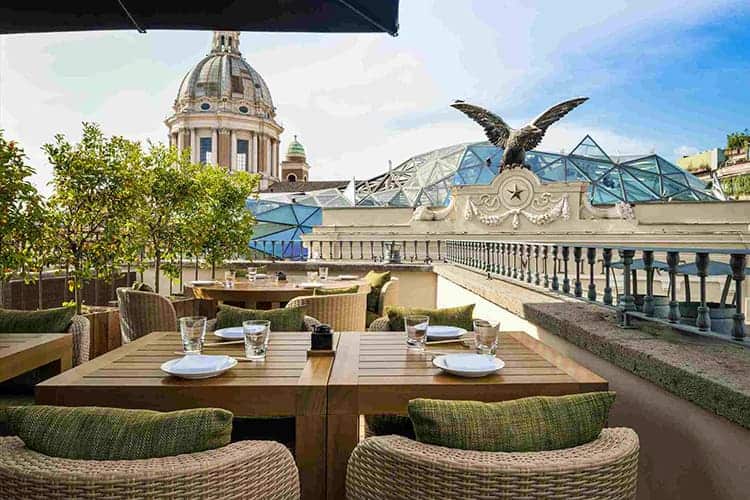 Elegant rooftop terrace in Rome with outdoor dining, classical architecture, green plants, and iconic historic buildings under a clear blue sky.