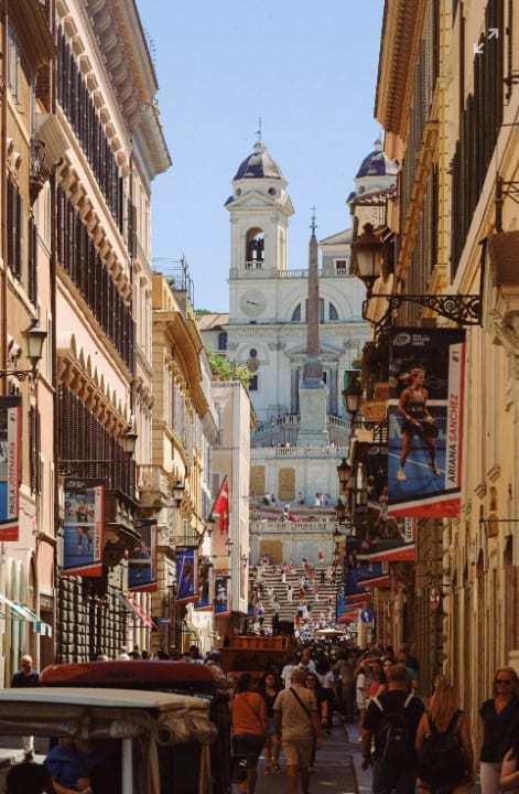 Vibrant street scene in Rome featuring historic buildings, bustling pedestrians, and the iconic Spanish Steps in the background under a clear blue sky.