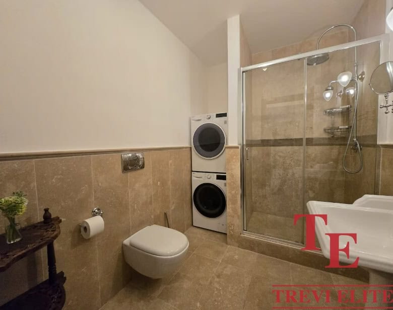 Luxurious modern bathroom with a glass-enclosed shower, wall-mounted toilet, and laundry appliances, featuring elegant beige tiles and sophisticated fixtures for a contemporary Italian home.