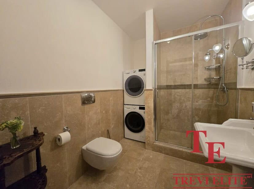 Luxurious modern bathroom with a glass-enclosed shower, wall-mounted toilet, and laundry appliances, featuring elegant beige tiles and sophisticated fixtures for a contemporary Italian home.