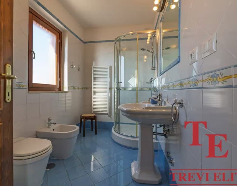 Modern bathroom with blue tiled floor, glass shower enclosure, white sink, bidet, and window providing natural light, in an elegant Italian property listed by Trevi Elite.
