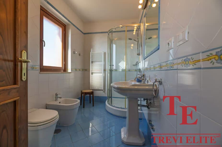 Modern bathroom with blue tiled floor, glass shower enclosure, white sink, bidet, and window providing natural light, in an elegant Italian property listed by Trevi Elite.