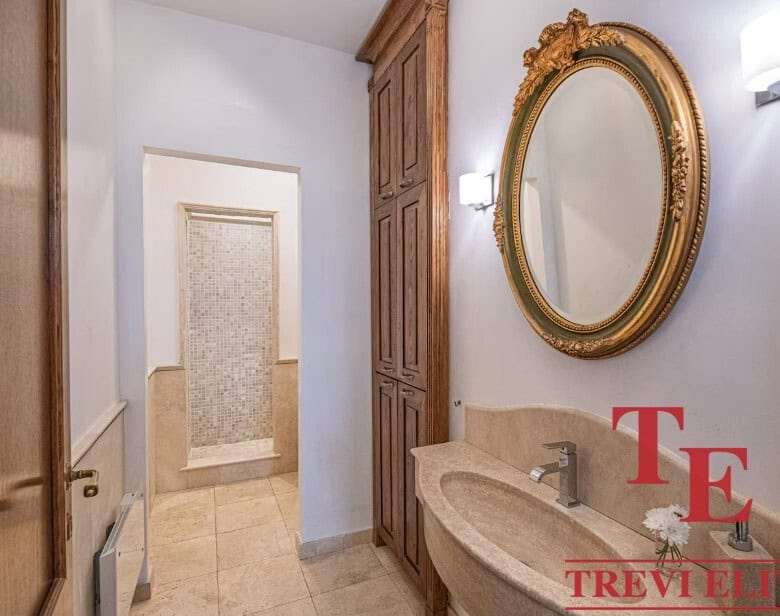 Elegant bathroom interior with a gold-framed mirror, double sink, wooden cabinet, marble countertop, and modern lighting, showcasing luxurious Italian real estate by Trevi Elite.