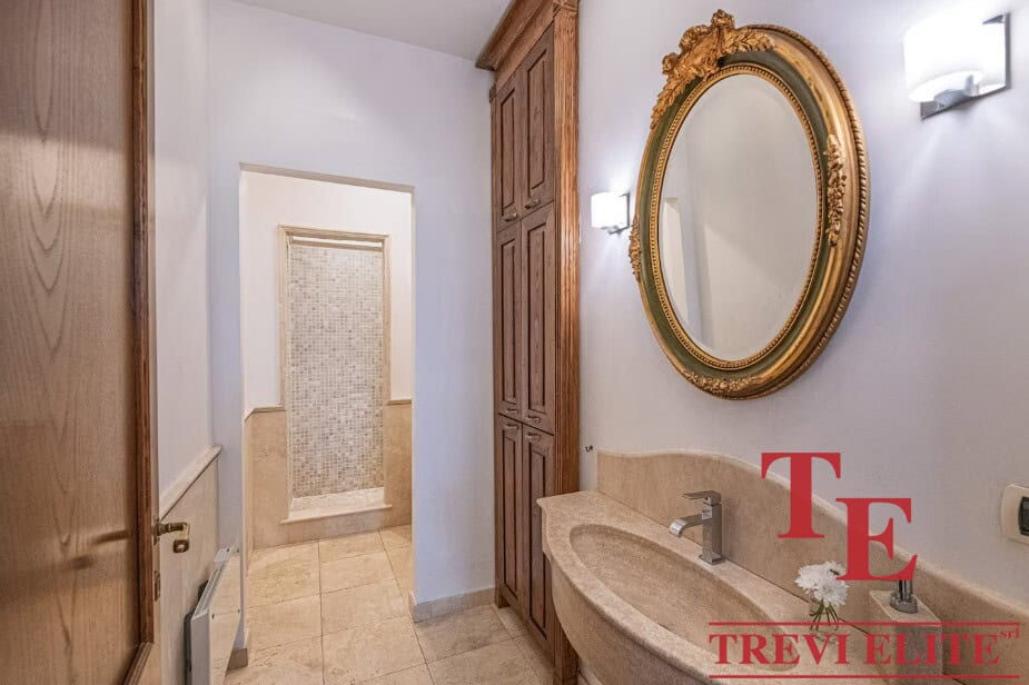Elegant bathroom interior with a gold-framed mirror, double sink, wooden cabinet, marble countertop, and modern lighting, showcasing luxurious Italian real estate by Trevi Elite.