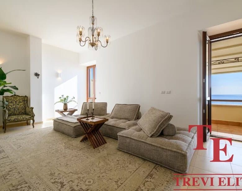 Bright living room with large windows and access to a balcony overlooking the sea, elegant chandelier, classic decor, and natural light creating a cosy, inviting atmosphere.