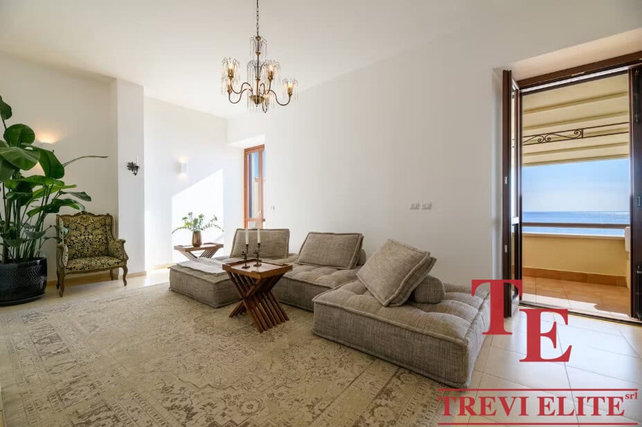 Bright living room with large windows and access to a balcony overlooking the sea, elegant chandelier, classic decor, and natural light creating a cosy, inviting atmosphere.