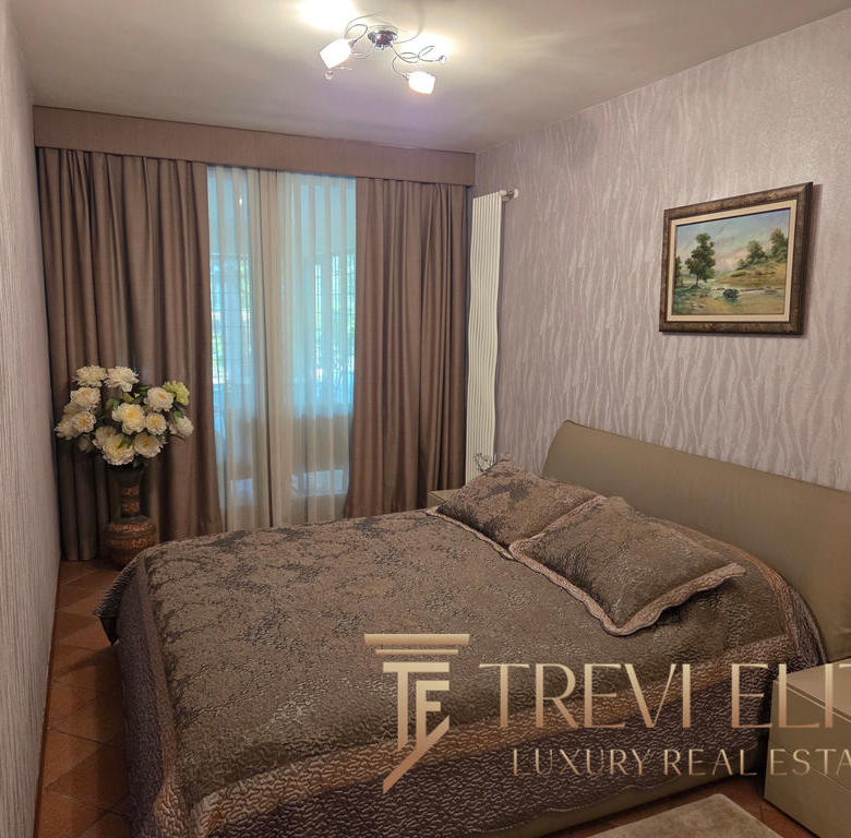Elegant bedroom with neutral tones, plush bedding, large window with beige curtains, framed landscape artwork, modern lighting, and decorative flowers, showcasing luxurious Italian real estate by Trevi Elite.