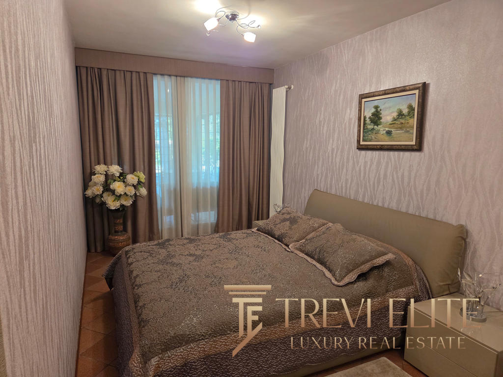 Elegant bedroom with neutral tones, plush bedding, large window with beige curtains, framed landscape artwork, modern lighting, and decorative flowers, showcasing luxurious Italian real estate by Trevi Elite.