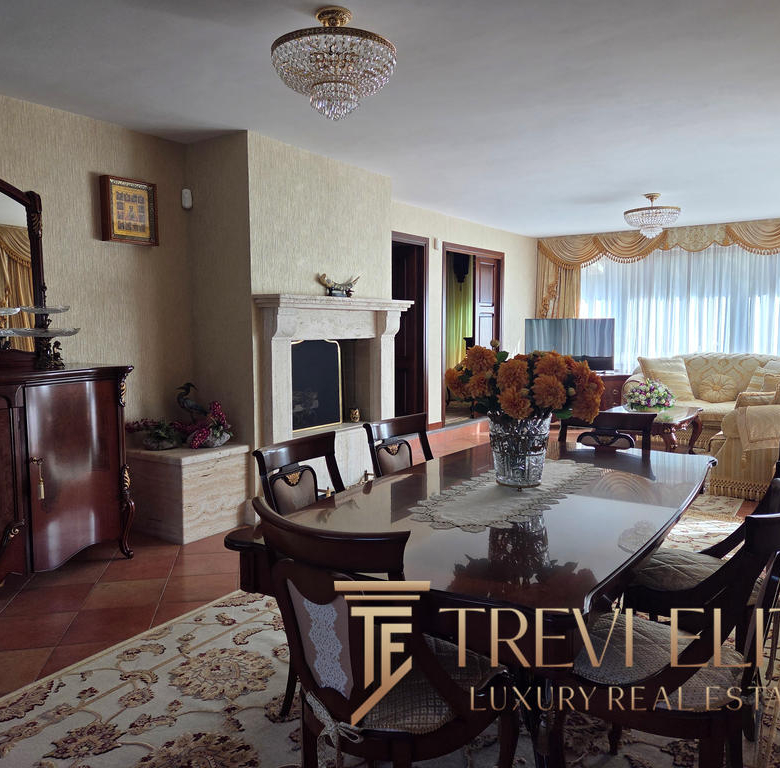 Elegant living room with classic furnishings, a large fireplace, floral arrangements, and a spacious window with gold drapes, showcasing luxurious Italian real estate by Trevi Elite.