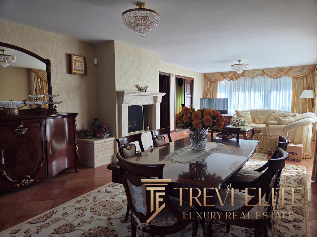 Elegant living room with classic furnishings, a large fireplace, floral arrangements, and a spacious window with gold drapes, showcasing luxurious Italian real estate by Trevi Elite.